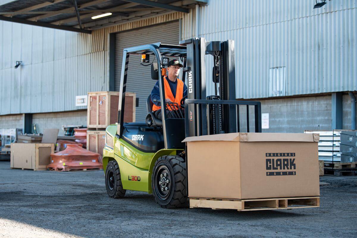 Man operating a Clark forklift to transport heavy equipment boxes within an industrial or warehouse setting.
