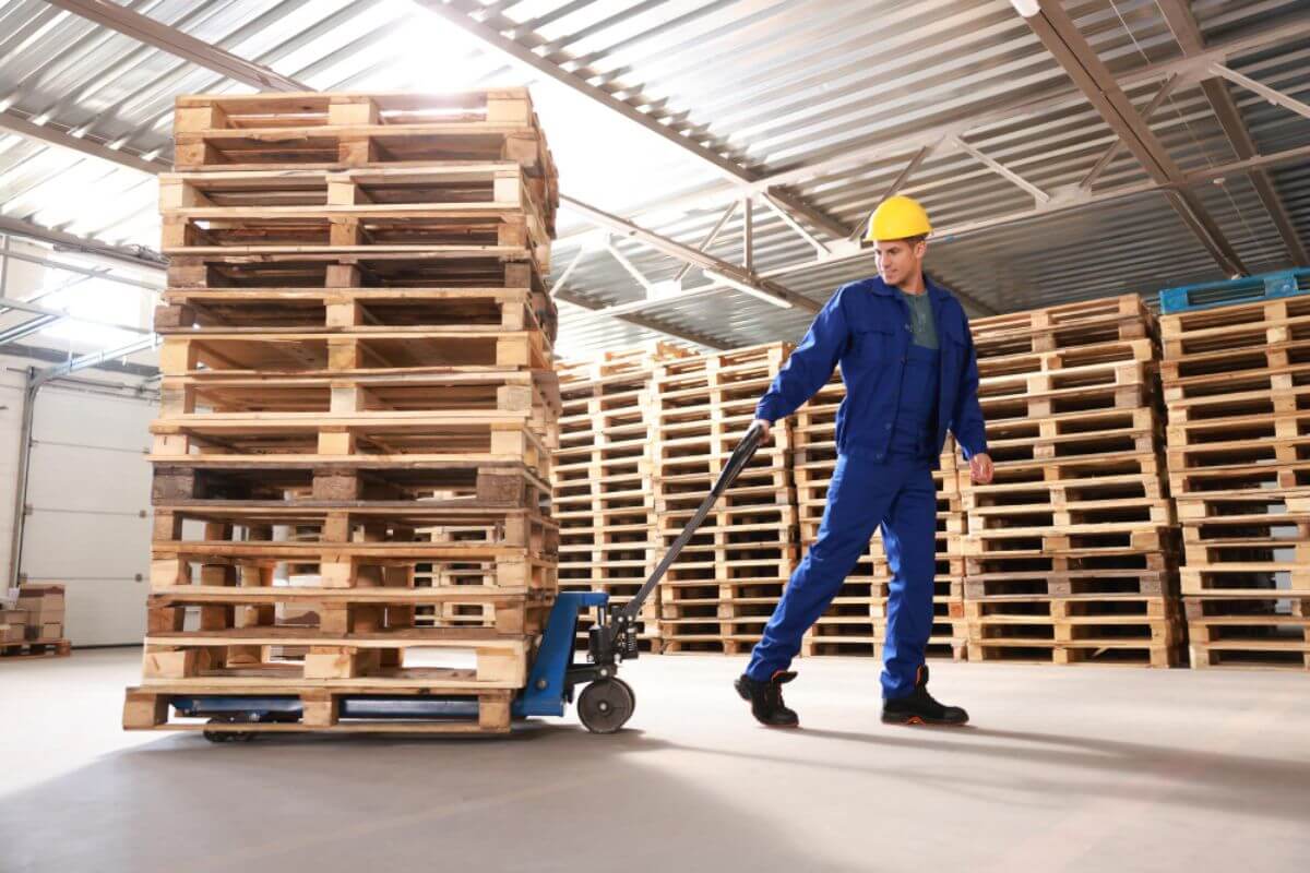 Factory worker operating a pallet truck to transport heavy wood materials within an industrial production environment.