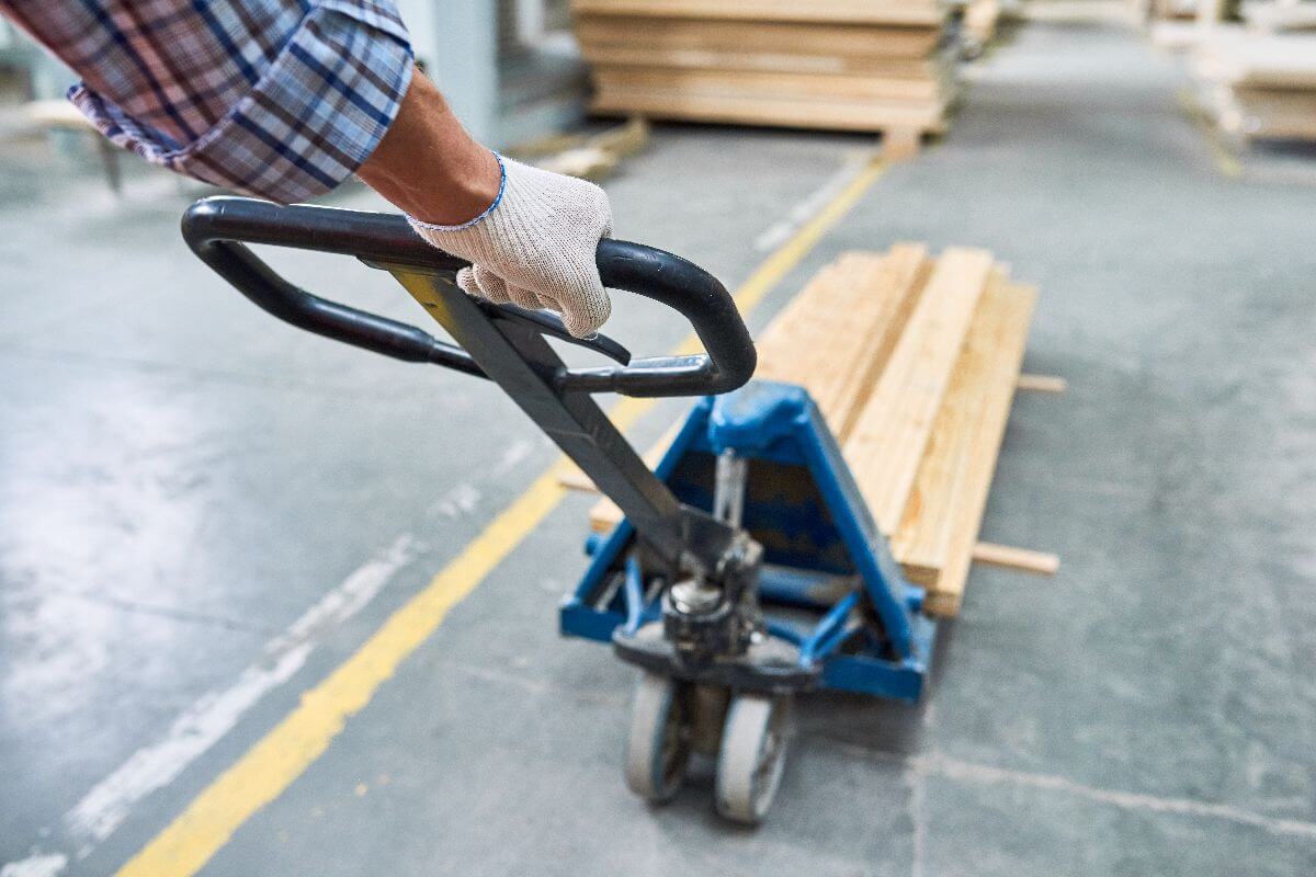 Man pulling a hand pallet truck while transporting wood panels, demonstrating efficient material handling in an industrial workspace.