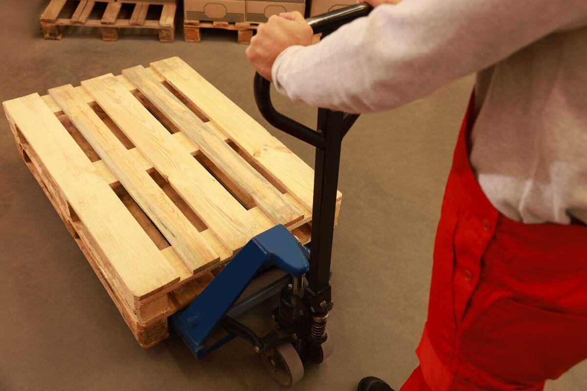 Warehouse worker using a blue hand pallet truck to transport wood materials efficiently within an industrial storage facility.