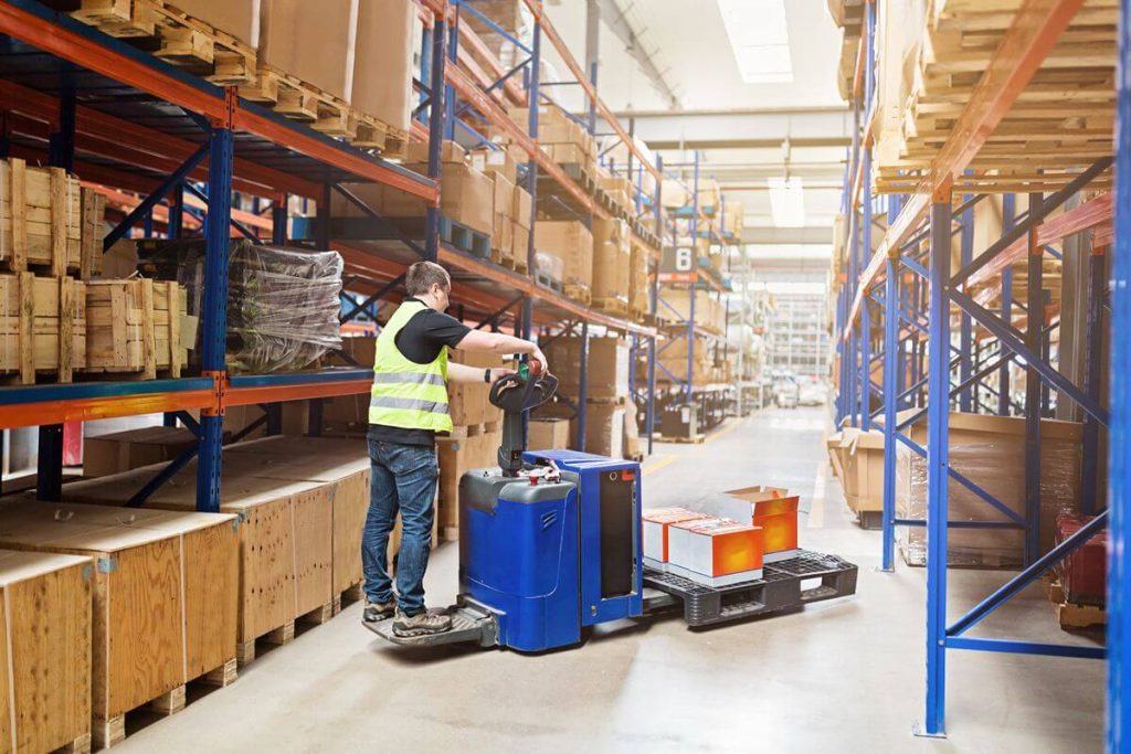 Warehouse operator using an electric pallet truck to move a pallet load through a busy storage aisle